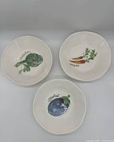 Top view of the set of three ceramic bowls with painted vegetables: artichoke, carrots with herbs, eggplant, on white background.