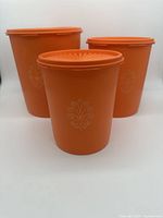 Three orange plastic Tupperware nesting canisters with lids, arranged side-by-side with a plain white background. Each canister has a decorative embossed flower pattern on the front.