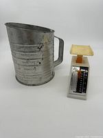 Photo showing metal flour sifter with embossed cup measurements and handle alongside small diet scale with cream platform on a white background