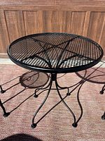 Round black wrought iron mesh top bistro table with star pattern reinforcement underneath and curved legs, placed on outdoor mat.