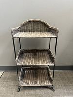Frontal view of the 3-tier shelf showing top curved wicker shelf and three woven shelves supported by wrought iron legs on carpeted floor.