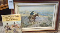 Framed painting of a cowboy on horse with Western landscape by Charles M. Russell, shown alongside the book with cowboy illustration cover
