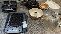 Assorted bakeware items including muffin tins, bunt pans, fry pans, racks seen on carpeted floor.