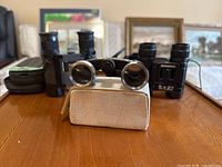 Front view of three optical items on a wooden surface; Mignon opera glasses with case in front, two Bushnell binoculars behind.