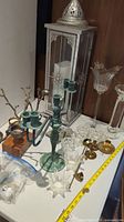 Photo showing tall silver metal and glass lantern, green metal candelabra, clear glass candle holders, brass and branch-style candle holders on white table
