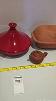 Shows three cookware items: a red tagine, a brown clay roaster, and a small Japanese-style teapot next to a measuring tape for size reference.