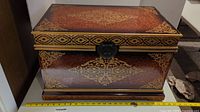 Front view of a rectangular decorative box with dark reddish-brown lacquer and gold ornamental floral and geometric patterns, showing a metal latch closure and some wear marks.
