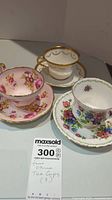 Three fine china teacups each with matching saucers, featuring different floral patterns and gold trim, arranged on a table with a Maxsold note showing quantity and description.