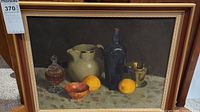 Full view of framed still life painting showing jug, bottle, glass container, small bowl, brass cup, and oranges on patterned cloth.