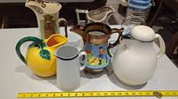 Set of five jugs of various styles, sizes, and materials placed on counter with measuring tape showing sizes between about 7 and 10.5 inches.