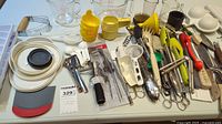 Overview photo showing diverse kitchen utensils including metal and plastic peelers, tongs, graters, measuring cups, white plastic lids, and scoops.