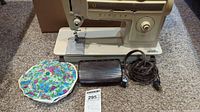 Singer sewing machine with foot pedal, electrical cord, and floral pincushion accessory on carpeted floor.