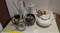Wide shot showing six assorted tea and coffee pots of varying sizes and materials on a white shelf. Includes white ceramic pot missing lid, colorful patterned pot, tall white pot, painted metal pot, and silver metal pot.