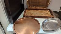 Photo showing four trays: wooden inlay tray with detailed pattern, wicker tray behind it, round copper tray in front left, and smaller oval metal tray on front right.