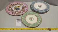 Overview of multiple plates and platters with varied floral and ornate border designs, including pink platter, blue-bordered plates, and green swirl patterned plates.