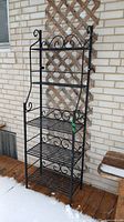 Full front view of the black wrought iron baker's rack showing five slatted shelves and decorative curly cue details against a brick wall outdoors