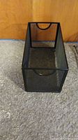 Tall narrow black mesh metal document organizer sitting upright on carpeted floor in front of wooden backdrop