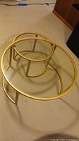 Two round nesting coffee tables with matte gold frames and clear glass tops, arranged nested together on carpeted floor.