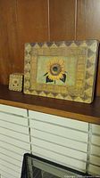 Photo showing one rectangular placemat and one square coaster, both featuring sunflower design with geometric borders, placed on wooden mantle.