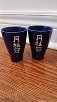 Two blue ceramic saki cups with white Asian characters, tapered design, on wood floor near white baseboard