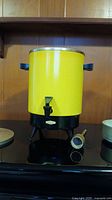 Full view of large yellow vintage Empire coffee urn with black base, black handles, and spigot. Metal filter, stem and lid placed beside it.