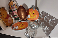Nine cake pans arranged on a shelf including character shaped pans of Winnie the Pooh, rag doll, football, two pumpkins with jack-o'-lantern faces, a Halloween pumpkin pan with five smaller pumpkins, and a young girl/doll pan.
