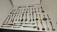 Photo showing the full collection of antique forks, spoons, butter spreaders and ladles arranged on white surface.
