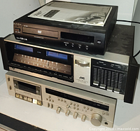Stacked view of all three electronic devices: Apex DVD player on top, JVC receiver in middle, Panasonic cassette deck on bottom, with paperwork on top.