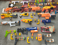 Overview of the large assortment of Nerf blasters laid out on floor including various colors and sizes, numerous clip magazines and accessories, plus a storage bin.