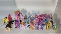 Photo showing a large collection of My Little Pony toys arranged on a white backdrop. Different sizes and colors represented including some large ponies with brushable hair and smaller collectible figures.