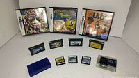 The photo shows three Nintendo DS game cases with visible cover art and various loose Nintendo DS game cartridges arranged in front, including a blue plastic carrying case.