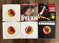 Top view of six Bob Dylan 45rpm singles laid out, showing three singles in white paper sleeves, one in black Columbia sleeve, and two cardboard printed sleeves featuring Bob Dylan imagery.