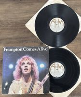 Two vinyl records and one album cover of Peter Frampton's 'Frampton Comes Alive' shown with one record out of sleeve and the other partially visible.