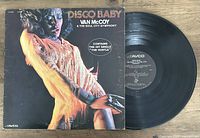 Photo of the Van McCoy 'Disco Baby' vinyl record and album cover showing front cover with a woman dancing in disco attire. Vinyl record is partially removed from the sleeve to show label.