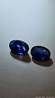 Pair of natural oval cut blue sapphire gemstones on light surface under natural light, showing deep blue color and faceting.