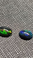 Close-up image of two natural oval black opals on a textured black surface, showing bright multicolored flashes of green, blue, orange, and red.