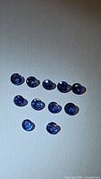 Eleven loose natural round cut blue sapphires positioned on a light surface, displaying faceted round cuts with a deep blue color.