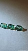 Three natural octagon cut emerald gemstones arranged side by side on a light surface, showing their green color and visible inclusions