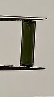 Close-up of rectangular baguette cut green tourmaline held by tweezers, showing color and clarity.