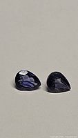 Pair of natural deep purple pear cut iolite gemstones resting on a neutral surface showing facets and color.