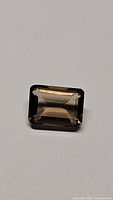 Top-down view of rectangular emerald cut smokey quartz gemstone on a plain surface showing clarity and color.