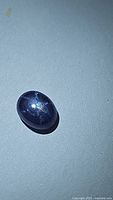 Oval cabochon natural star sapphire with visible 6-ray star reflection under focused torch light on a plain background.