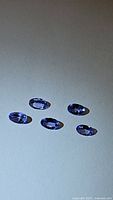 Five calibrated faceted oval tanzanite gemstones with natural blue-purple color placed on a white surface showing clarity and color.