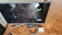 Samsung TV front view showing screen, silver stand, and cables on floor including Apple TV and power adapter