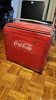 Front view of red Coca-Cola metal cooler with white script logo, handle on top, and visible wear and rust.