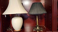 Two table lamps side by side on shelf. One brass lamp with black shade, one ceramic crackle glazed lamp with white shade and wooden broken base.