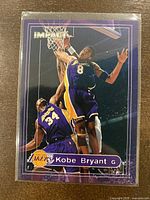 Front of 1999-00 Fleer Impact Kobe Bryant basketball card showing Kobe Bryant (#8) in Lakers uniform jumping near the basket with Shaquille O'Neal (#34) in the background, with card title and branding.