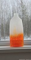 Tall rectangular art glass vase with frosted white top and orange gradient bottom placed on a snow-covered window sill, showing texture and coloration.
