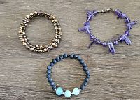 Three bracelets displayed on a flat surface including a bronze-coloured pearl coil, purple quartz bead bracelet with clasp, and black lava bead with aquamarine bead stretchy bracelet.