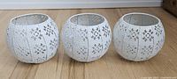 Three white tin punched candle holders with floral cutout pattern seen on wooden floor.